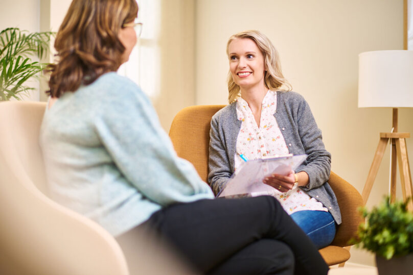 naisasiakas ja naispsykoterapeutti istuvat vastakkain kun hymyilee ja täyttää asioita paperille
