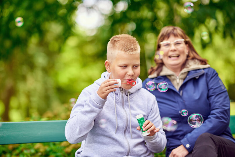 poika puhaltaa saippuakuplia puistonpenkillä ja vieressään istuva nainen nauraa