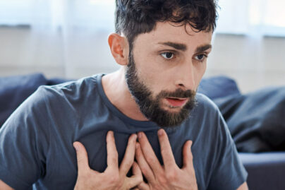 Parrakas mies istuu kotona lattialla ja pitää käsiä rinnallaan paniikkikohtauksen aikana.