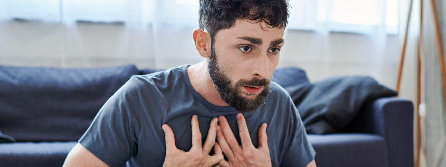 Parrakas mies istuu kotona lattialla ja pitää käsiä rinnallaan paniikkikohtauksen aikana.