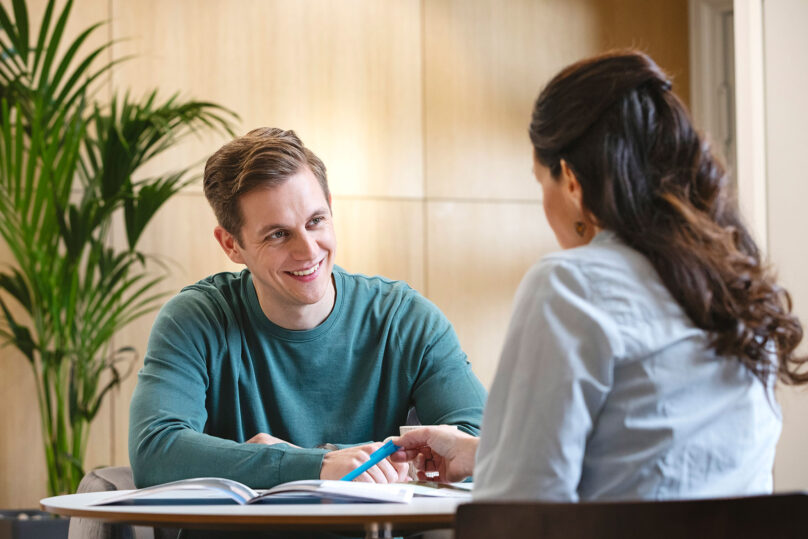 Naispsykologi ja nuori hymyilevä miesasiakas keskustelevat pöydän ääressä, psykologilla kynä kädessä.