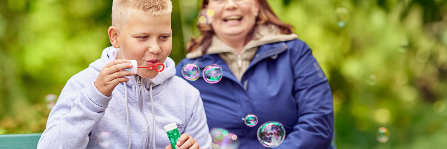 Poika puhaltaa saippuakuplia puistonpenkillä ja vieressä istuva nainen nauraa.