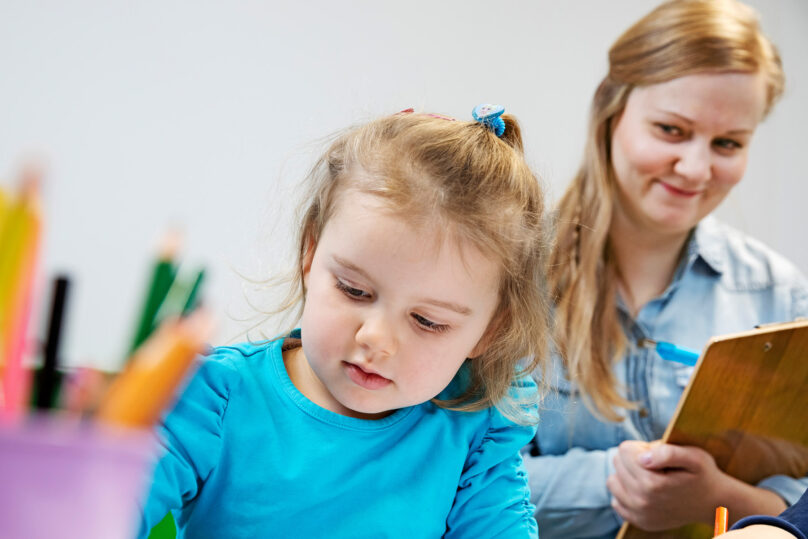 Lähikuvassa lapsi piirtämässä ja taustalla psykologi kirjaa merkintöjä paperille.