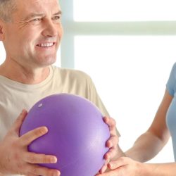mies jumppaa pallon avulla ja fysioterapeutti ohjaa vieressa