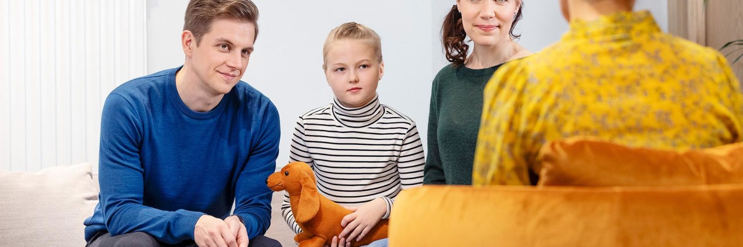Isä, tyttölapsi ja äiti istuvat sohvalla perheterapiassa ja katsovat terapeuttia.