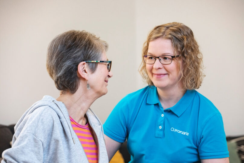 Harmaahiuksinen nainen ja kiharahiuksinen puheterapeutti hymyilevät toisilleen.