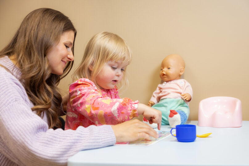 Tummahiuksinen puheterapeutti ja vaaleahiuksinen tyttölapsi tekevät pöydän ääressä harjoitusta vauvanuken kanssa.
