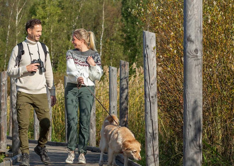 Mies ja nainen kävelemässä pitkospuilla, taluttaen koiraa virkeän ja hyvin nukkuneen näköisinä.