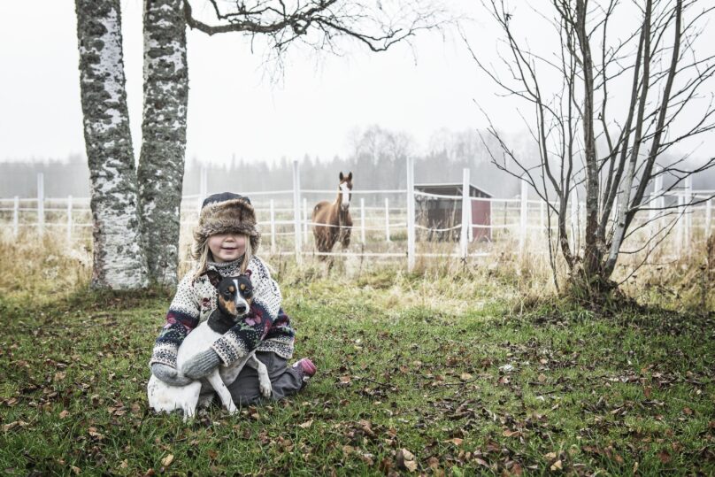 Tyttö istuu maassa koira sylissä ja taustalla on syksyinen hevostila.