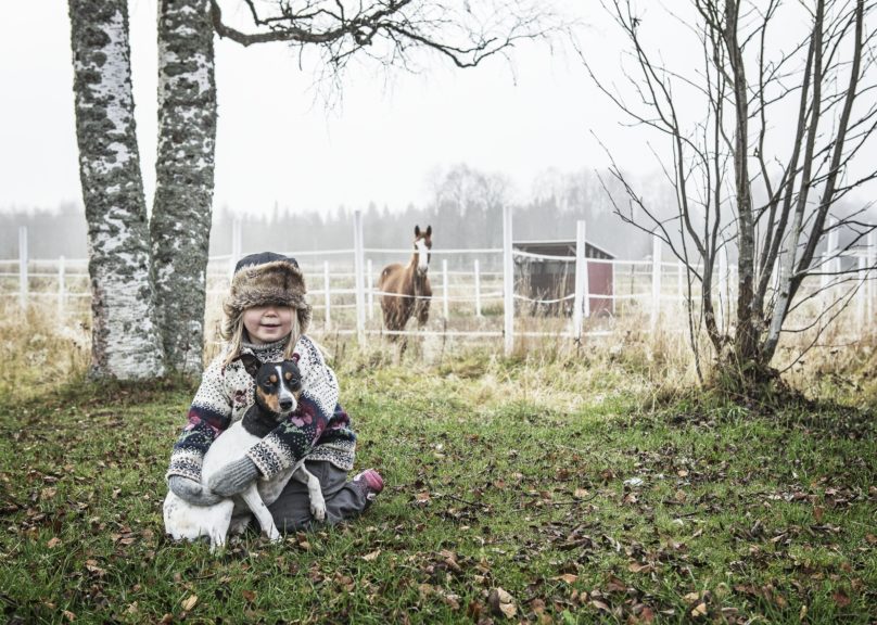 Tyttö istuu maassa koira sylissä ja taustalla on syksyinen hevostila.