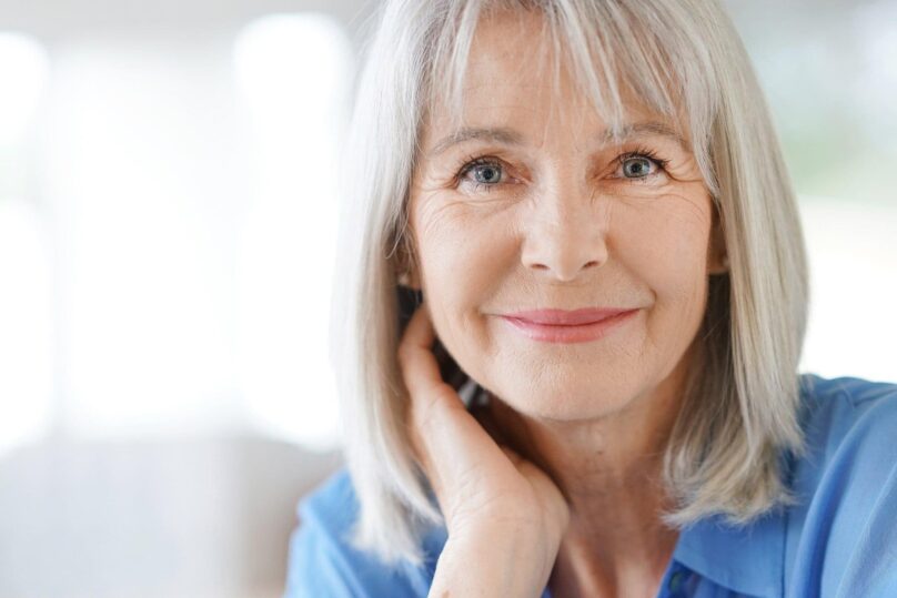 Vaaleahiuksinen iäkkäämpi nainen katsoo kohti ja hymyilee hienoisesti