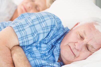 Iäkäs, valkohiuksinen mies nukkuu tyytyväisenä sängyssä.