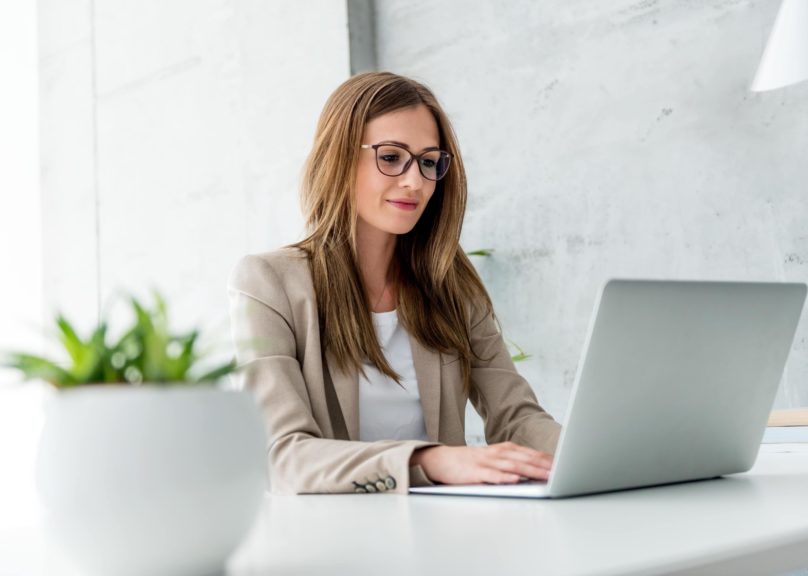 Silmälasipäinen nainen istuu puku päällä tietokoneella.