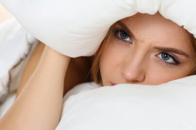 Nuori nainen tyynyn ja peiton välissä, sekä katsoo sieltä.