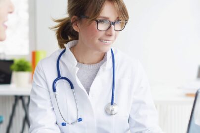 Ruskeahiuksinen naislääkäri ja naispotilas istuvat pöydän ääressä lääkärin käyttäessä kannettavaa tietokonetta.