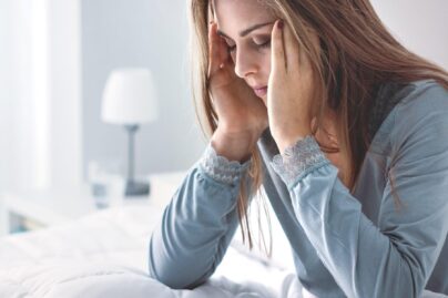 Tummahiuksinen nainen istuu väsyneenä sängyllä kädet poskilla.