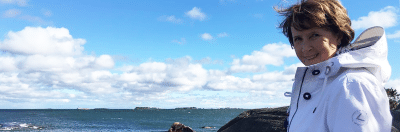 Nainen hymyilee ja katsoo kameraan ulkona veden äärellä.