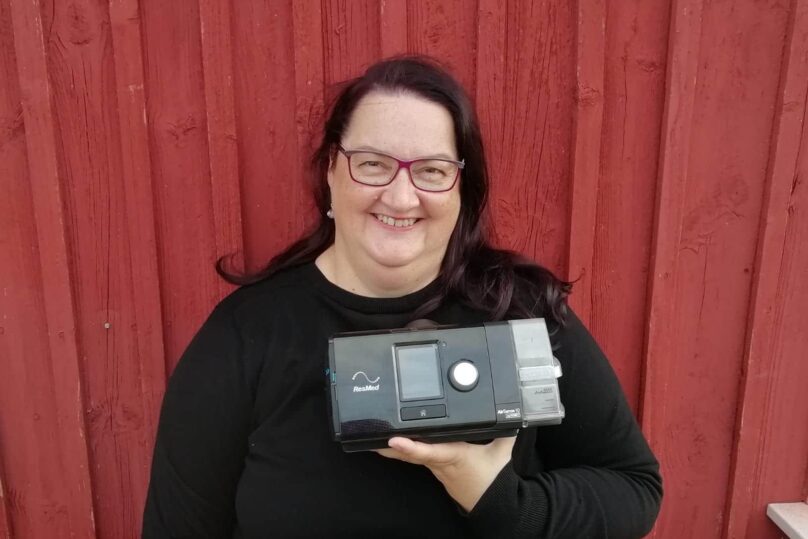Eija Sjöblom CPAP-laitteensa kanssa punaisen seinän edessä