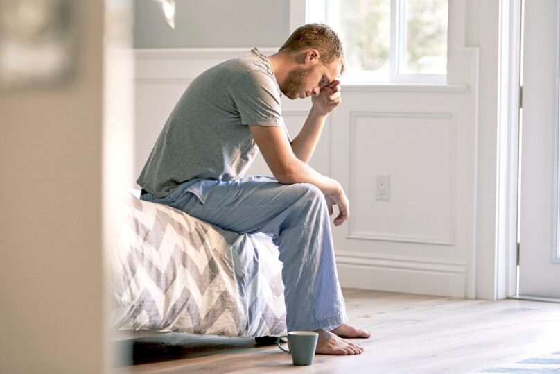 väsynyt mies nojaa käteensä sängyn reunalla