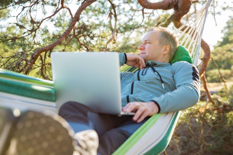mies makoilee riippukeinussa ja nojaa kateen kannettava tietokone sylissa
