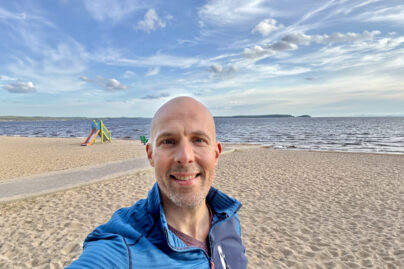 Aikuinen mies ottaa selfien hiekkarannalla, taustalla meri ja sininen taivas pilvineen.