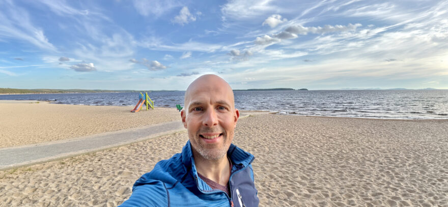 Aikuinen mies ottaa selfien hiekkarannalla, taustalla meri ja sininen taivas pilvineen.