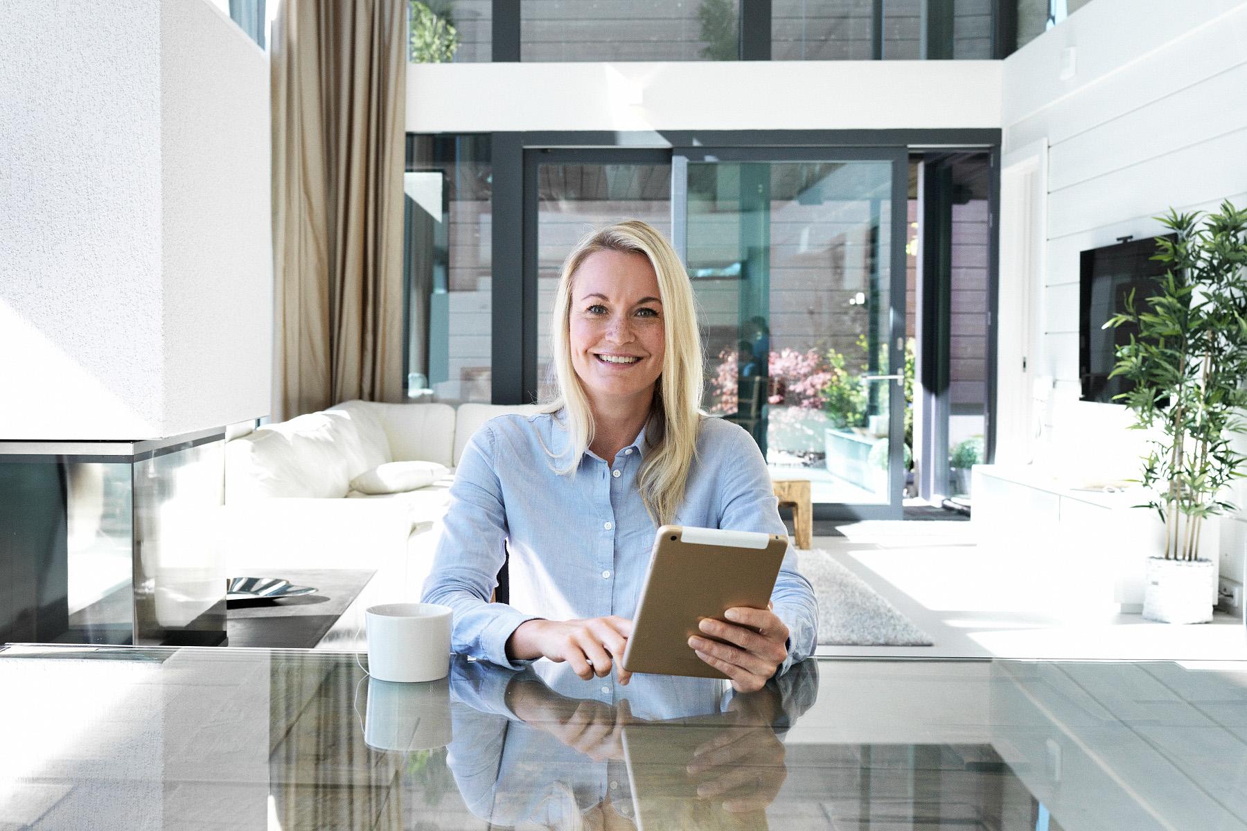 Vaaleahiuksinen nainen istuu modernissa talossa pöydän ääressä iPad kädessään.