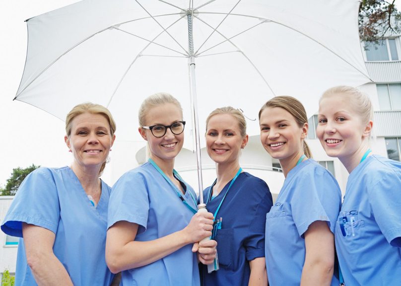 Viisi naispuolista hoitajaa seisoo ulkona sateenvarjon alla ja hymyilee.