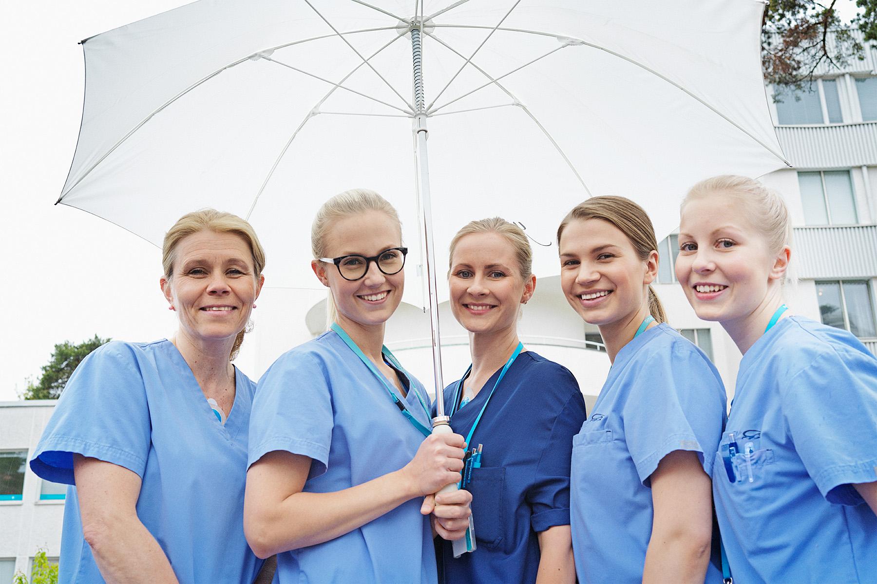 Viisi naispuolista hoitajaa seisoo ulkona sateenvarjon alla ja hymyilee.