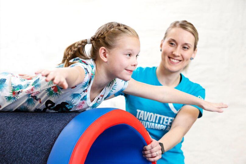 Coronaria kuntoutuksen toimintaterapeutti ja tyttöasiakas harjoittelee tasapainoa putken päällä maaten.