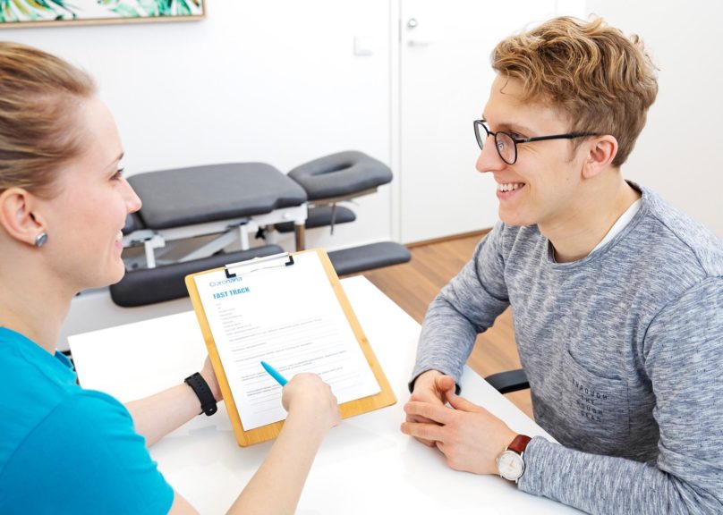 Kuntoutuksen naispuolinen fysioterapeutti ja miespuolinen asiakas istuvat pöydän ääressä ja nainen esittelee esitietolomaketta.