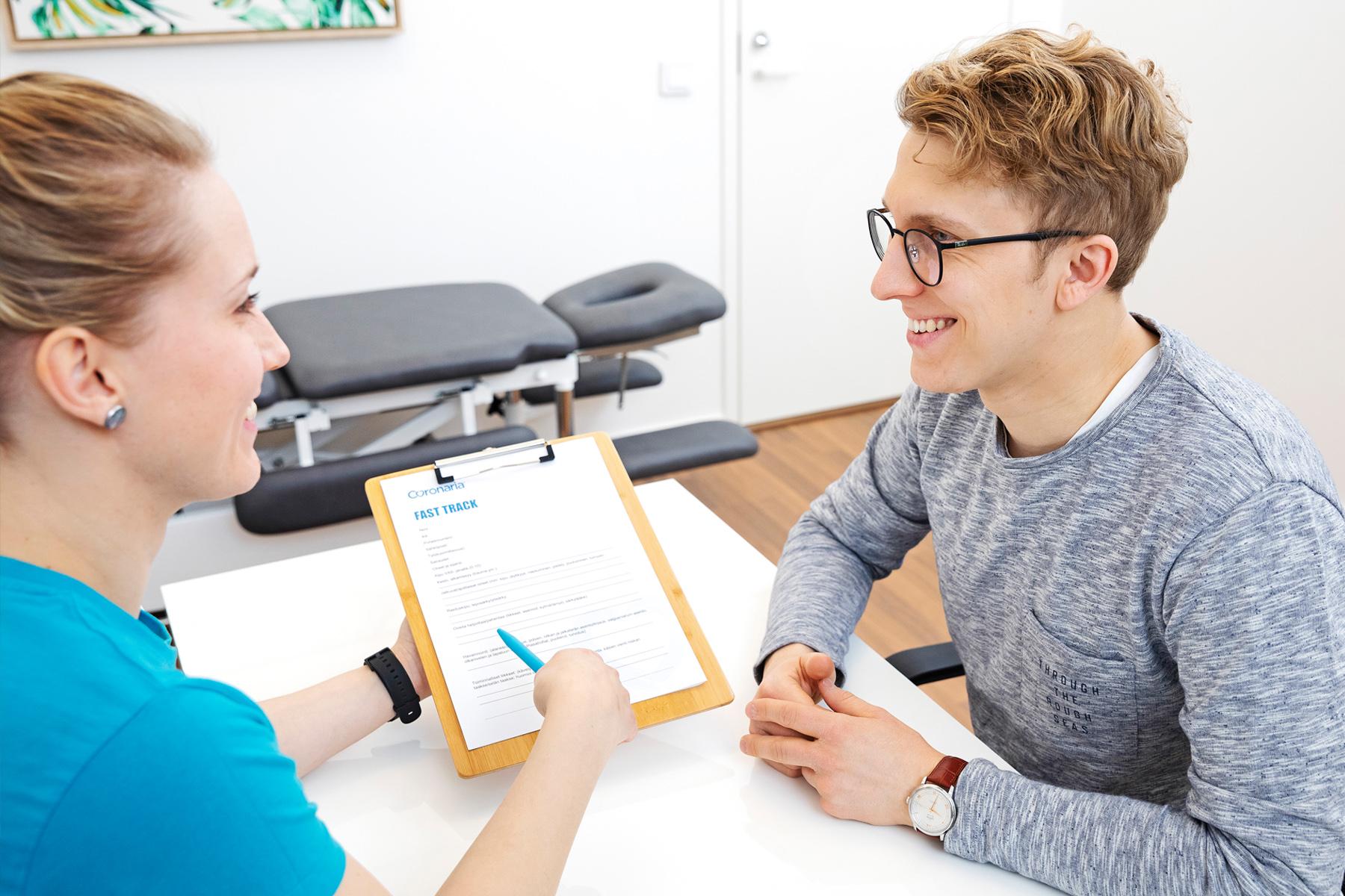 Kuntoutuksen naispuolinen fysioterapeutti ja miespuolinen asiakas istuvat pöydän ääressä ja nainen esittelee esitietolomaketta.