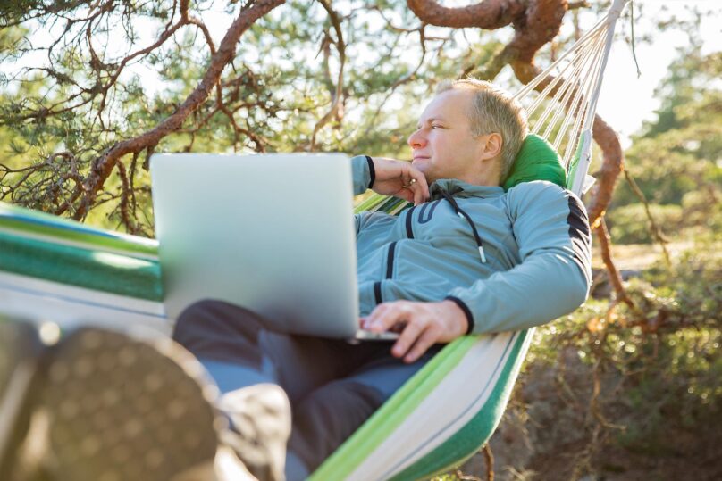 Mies makoilee riippukeinossa ulkona, läppäri sylissä.
