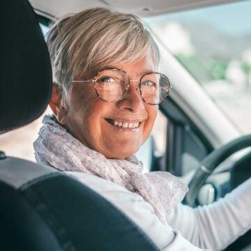 Seniorinainen auton ratissa katsoo kameraan hymyillen