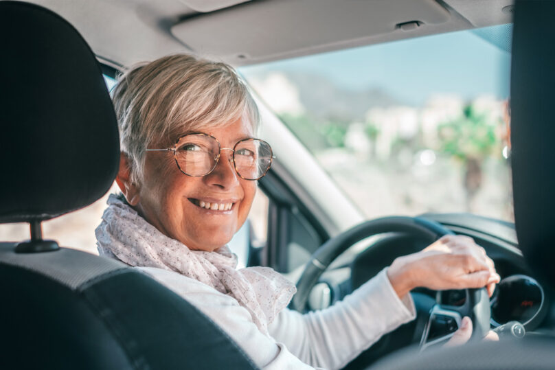 Seniorinainen auton ratissa katsoo hymyillen kameraan
