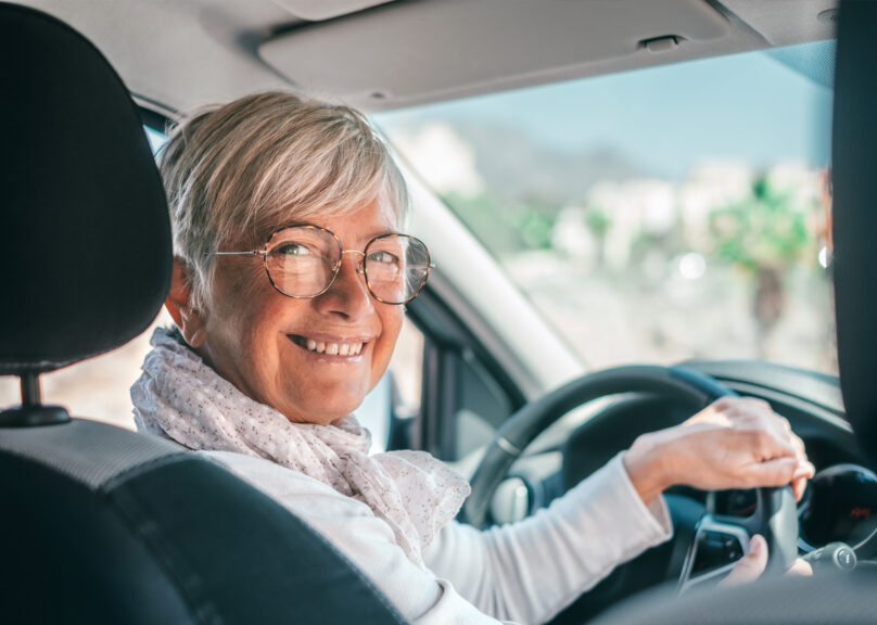 Seniorinainen auton ratissa katsoo hymyillen kameraan
