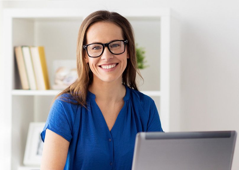 Hymyilevä nainen istuu tietokoneella ja katsoo kameraan.