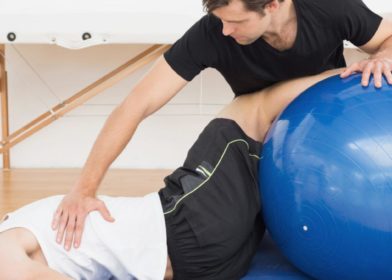 Fysioterapeutti auttaa nuorta miesasiakasta alaselkäkivussa jumppapallon avulla,