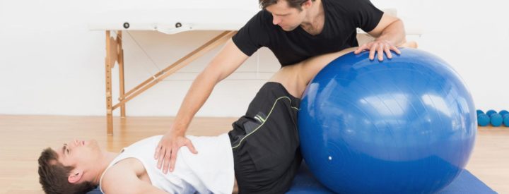Fysioterapeutti auttaa nuorta miesasiakasta alaselkäkivussa jumppapallon avulla,