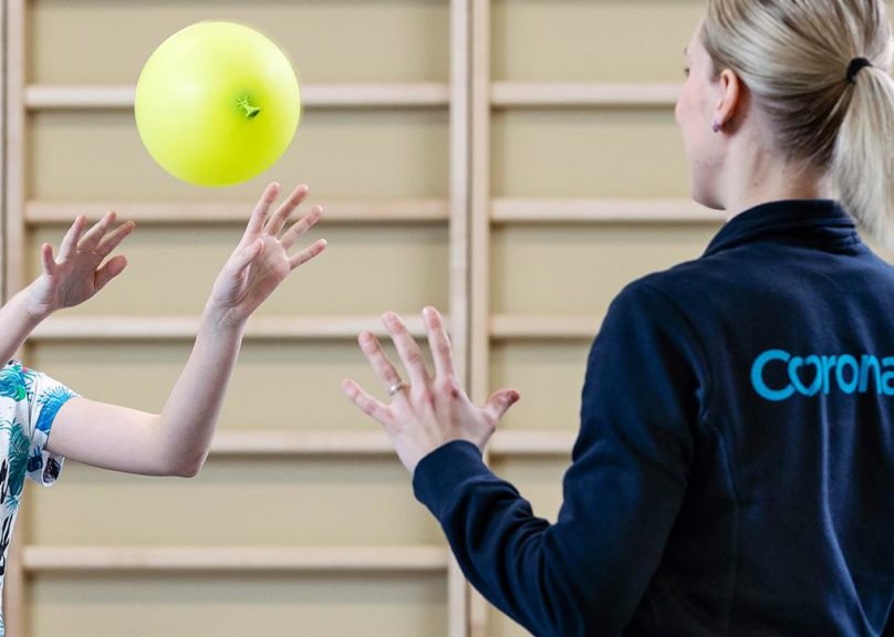 Coronarian fysioterapeutti ja hymyilevä lapsiasiakas heittelevät ilmapalloa fysioterapiassa.