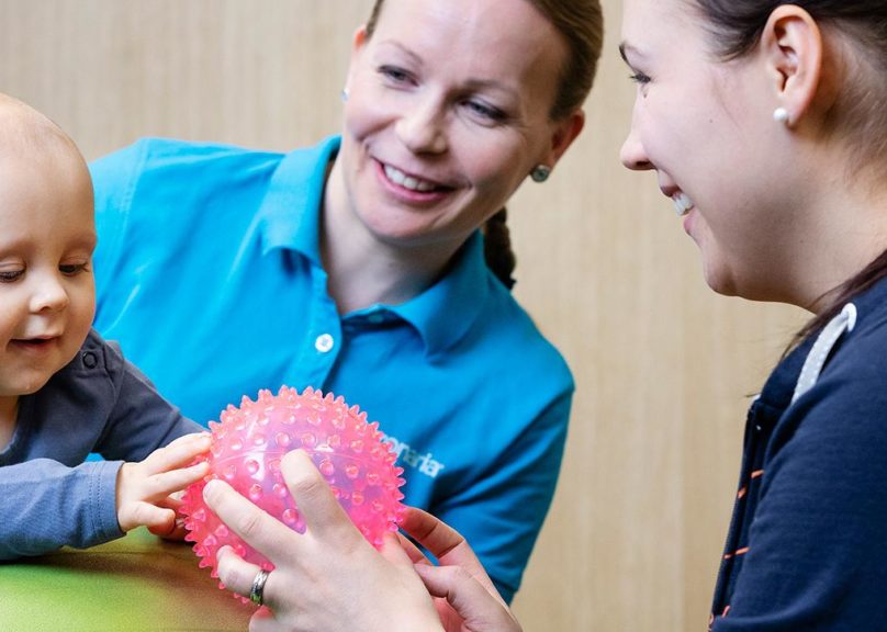 Vauva ja äiti leikkivät yhdessä pallolla äitiysfysioterapiassa ja Coronarian fysioterapeutti ohjaa vieressä liikkeitä.