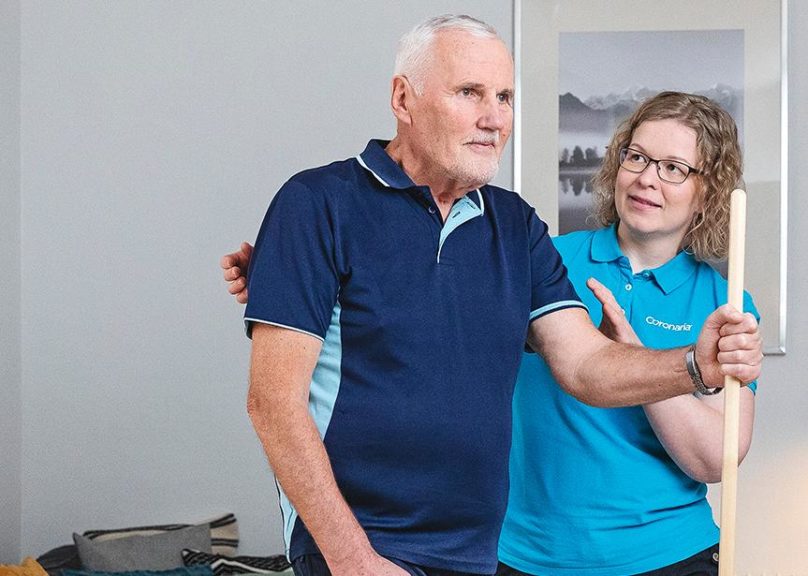 Seniorimiesasiakas keppijumppaa seisaaltaan ja fysioterapeutti ohjaa vieressä.