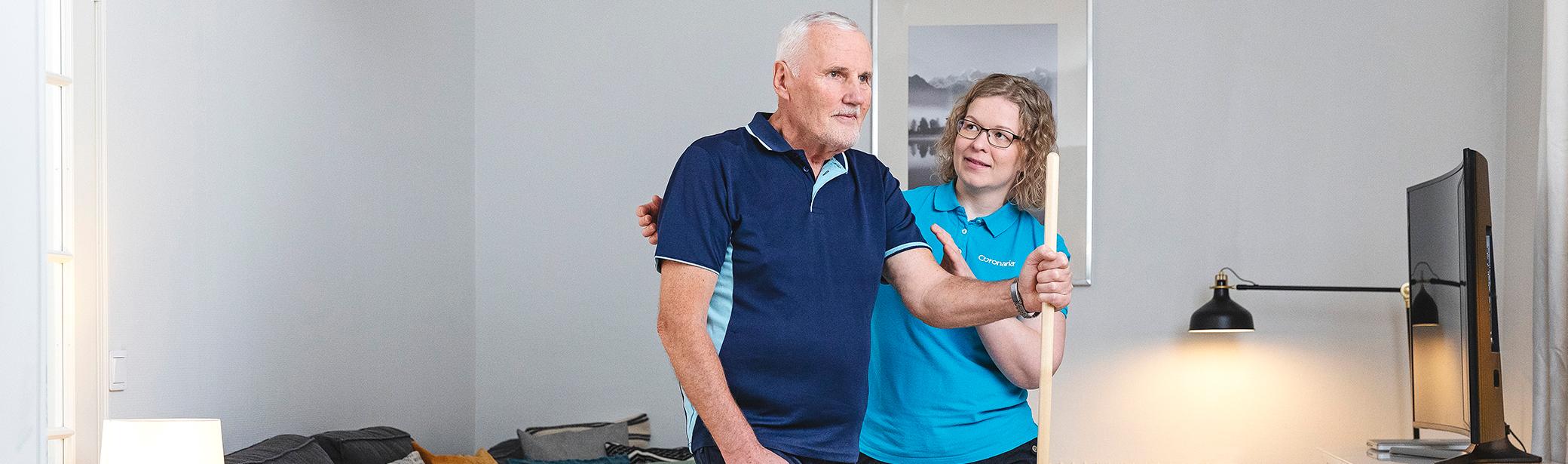 Seniorimiesasiakas keppijumppaa seisaaltaan ja fysioterapeutti ohjaa vieressä.