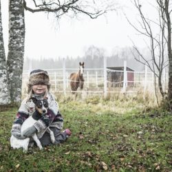 Tyttö istuu maassa koira sylissä ja taustalla on syksyinen hevostila.