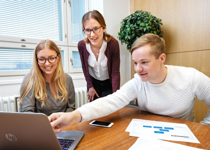 Kolme työntekjiää on kerääntynyt pöydän ääreen ja katsoo yhdessä kannettavan tietokoneen näyttöä.