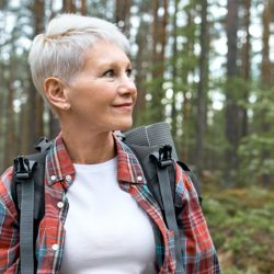 Harmaahiuksinen nainen kävelemässä metsässä, reppu selässä.