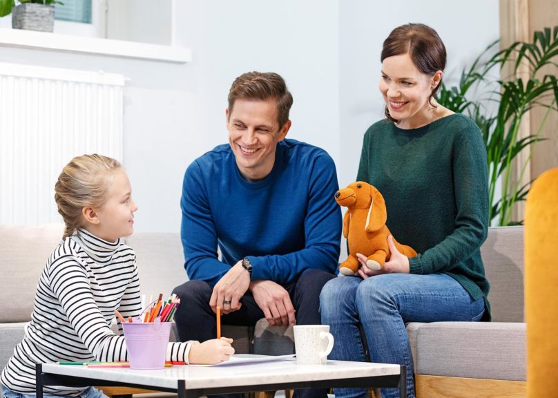 lapsi ja vanhemmat istuvat sohvaryhmassa ja hymyilevat