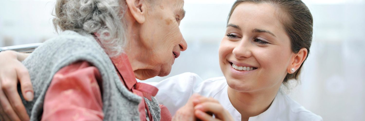 Nuori hoitajanainen ja vanha harmaahiuksinen nainen hymyilevät toisilleen.