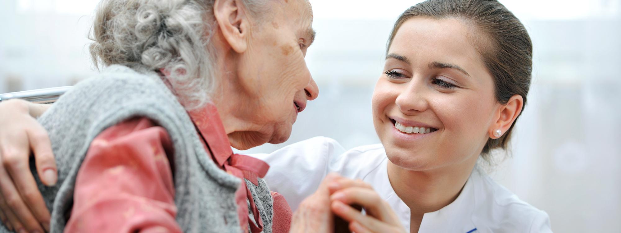 Nuori hoitajanainen ja vanha harmaahiuksinen nainen hymyilevät toisilleen.