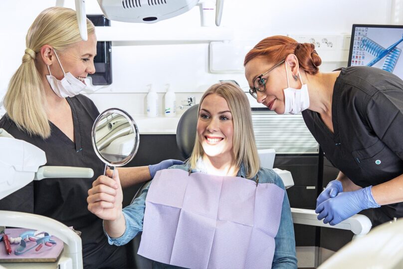 Työterveyshammashuollon naispuolinen asiakas katsoo ihaillen peiliin vierellään Coronarian hammaslääkäri ja hammashoitaja.